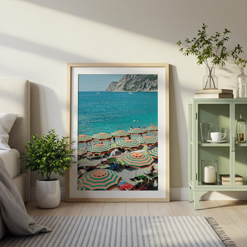 May include: A framed print of a beach scene with striped orange and green umbrellas. The turquoise water and a rocky cliff are visible in the background. The print is displayed in a light-colored wooden frame.