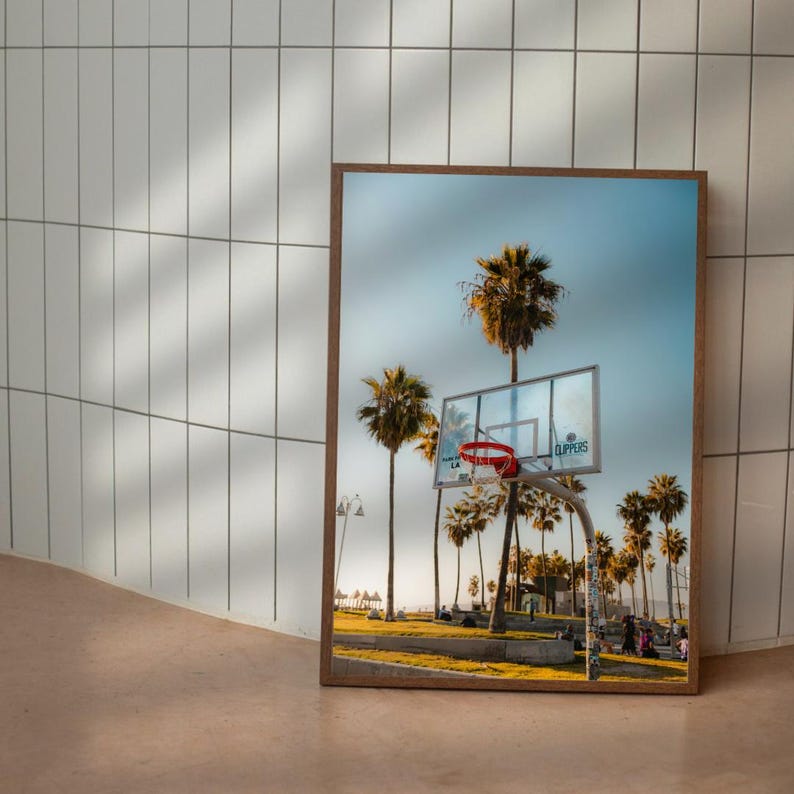 May include: A framed print showing a basketball hoop with a red net, set against palm trees and a blue sky. The image depicts a sunny day at a beachside court, with the word "CLIPPERS" visible on the backboard.