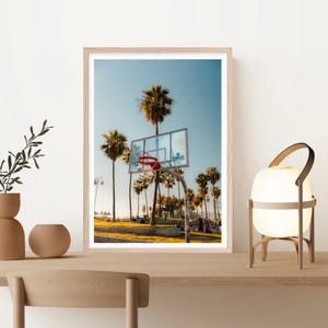 May include: A framed print featuring a basketball hoop with a red net, set against a backdrop of palm trees and a clear blue sky. The hoop is mounted on a metal pole, with the word "Clippers" visible on the backboard. The scene has a sunny, coastal feel.