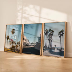 以下が含まれることがあります： カリフォルニア州ベニスビーチの風景を描いた、額装されたアートプリント3点。ヤシの木、バスケットボールコート、VENICEの文字が入った通り、スケートパークが描かれています。フレームは明るい木の色です。