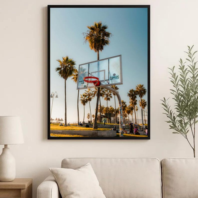 May include: A framed print showing a basketball hoop with a red net, set against palm trees and a blue sky. The image depicts a sunny day at a beachside court, with the word "Clippers" visible on the backboard. The scene evokes a sense of leisure.