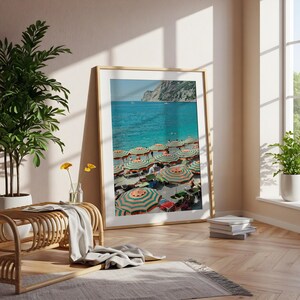 May include: A framed print of a beach scene with turquoise water and a multitude of striped umbrellas. The umbrellas are orange and green, providing shade for beachgoers. A mountain range is visible in the background.
