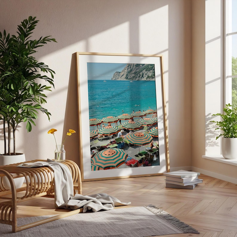 May include: A framed print of a beach scene with turquoise water and a multitude of striped umbrellas. The umbrellas are orange and green, providing shade for beachgoers. A mountain range is visible in the background.
