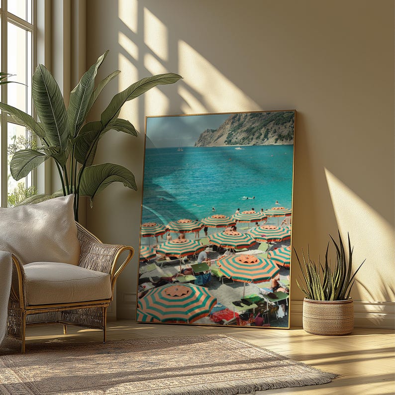 May include: A framed print of a beach scene with turquoise water, striped umbrellas, and a mountain in the background. The umbrellas have green and orange stripes. The print is displayed in a room with a wicker chair, a large plant, and a rug.