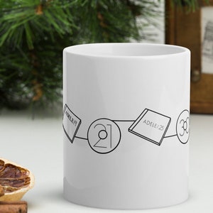 May include: White ceramic mug with a black line drawing of three album covers connected by a line. The album covers are "Adele 19", "21", and "Adele 25".