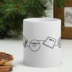 May include: White ceramic mug with a black line drawing of album art from the band Radiohead. The mug features the album titles "OK Computer" and "Kid A". The mug is a standard size for coffee or tea.