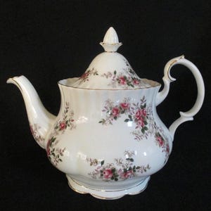 May include: A white porcelain teapot with a lid, decorated with pink roses and green leaves. The teapot features a curved spout, a loop handle, and gold trim. The design is a classic floral pattern.