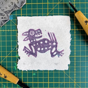 May include: A purple and white linocut print of a stylized creature on a white paper background. The print is on a green cutting mat.