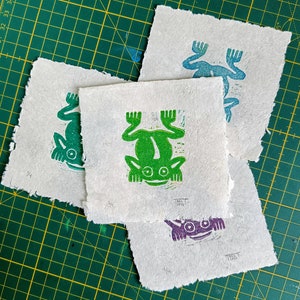May include: Four handmade paper cards with frog illustrations printed in green, blue, and purple ink. The cards are on a green cutting mat.
