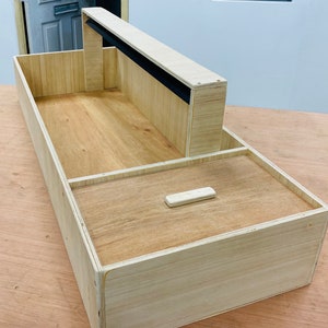 May include: A light-coloured wooden box with a raised section and a black strip. The box has a rectangular shape with a compartment and a small, rectangular wooden handle. The wood grain is visible.