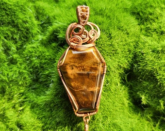 May include: A handcrafted pendant featuring a coffin-shaped tiger's eye stone, encased in copper wire. The pendant has a decorative wirework crown and a beaded dangle. The background is a bed of vibrant green moss.