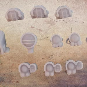 Puede incluir: Un surtido de pequeños objetos impresos en 3D de color gris claro. La colección incluye varias piezas en forma de gofre, una pequeña estructura rectangular, un objeto redondo con una línea horizontal y varias piezas con forma de elefante.