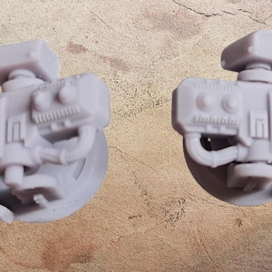 May include: Two light gray, futuristic-looking miniature gun models. Each model has a cylindrical barrel, a rectangular top piece, and intricate detailing. The models are set against a textured, brown background.