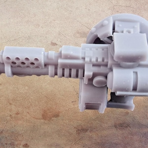 May include: A light gray, detailed miniature weapon, possibly a sci-fi or fantasy prop. The design includes a barrel with multiple holes, rectangular blocks, and a circular component. The object is set against a textured, brown background.