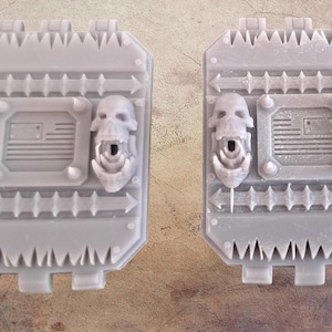 May include: Two gray, rectangular, mechanical-looking components. Each has a central panel with an American flag design, flanked by skull-shaped embellishments. The edges feature arrow and spike details, suggesting a futuristic or industrial theme.
