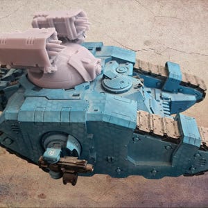 May include: A detailed model of a light blue tank with a large turret and two missile launchers. The tank has a textured surface and visible track treads. The turret and missile launchers are a light gray color.