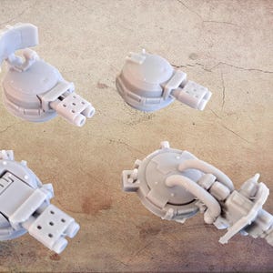 May include: Four light grey miniature turrets with detailed mechanical designs are arranged on a textured, tan surface. Each turret features a cylindrical body, multiple barrels, and intricate attachments, suggesting a futuristic or sci-fi theme.