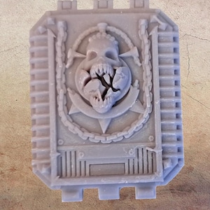 May include: A light gray rectangular object with a detailed skull design. The skull has a cracked jaw and is surrounded by a star, chains, and axes. The object has a textured surface and a series of vertical lines along the sides.