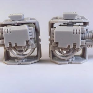 May include: Two gray, detailed miniature gun models, likely for tabletop gaming or model building. Each model features a complex design with a barrel, housing, and support structure. The models are displayed against a white background.