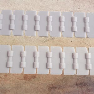 Puede incluir: Dos cadenas de plástico blanco, cada una compuesta por segmentos rectangulares unidos por bisagras cilíndricas. Las cadenas están dispuestas en paralelo, sobre un fondo neutro.