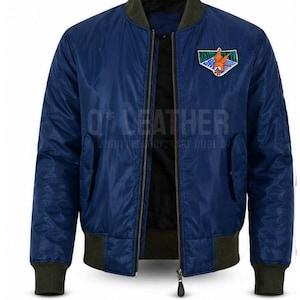 May include: A navy blue bomber jacket featuring a dark olive green ribbed collar, cuffs, and hem. The jacket has a front zipper closure and a colorful embroidered patch on the left chest depicting an orange character. The text Q*LEATHER is visible.