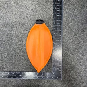 Puede incluir: Un objeto naranja en forma de lágrima con una parte superior negra. El objeto se coloca sobre una superficie gris junto a una regla. El objeto mide aproximadamente 25 cm de largo.