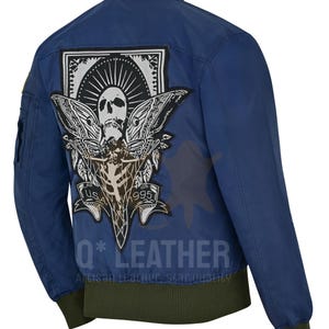 Puede incluir: Chaqueta bomber azul marino con un gran parche que presenta una calavera y alas. El cuello, los puños y el dobladillo son verdes. El texto "Q* LEATHER" está impreso en la parte inferior.