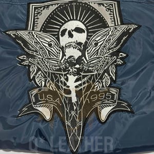 May include: A dark blue jacket featuring a large embroidered patch on the back. The patch displays a white skull with moth wings, a sunburst design, and a banner at the bottom with the text U.S.A. 1995. The embroidery uses black and white thread.