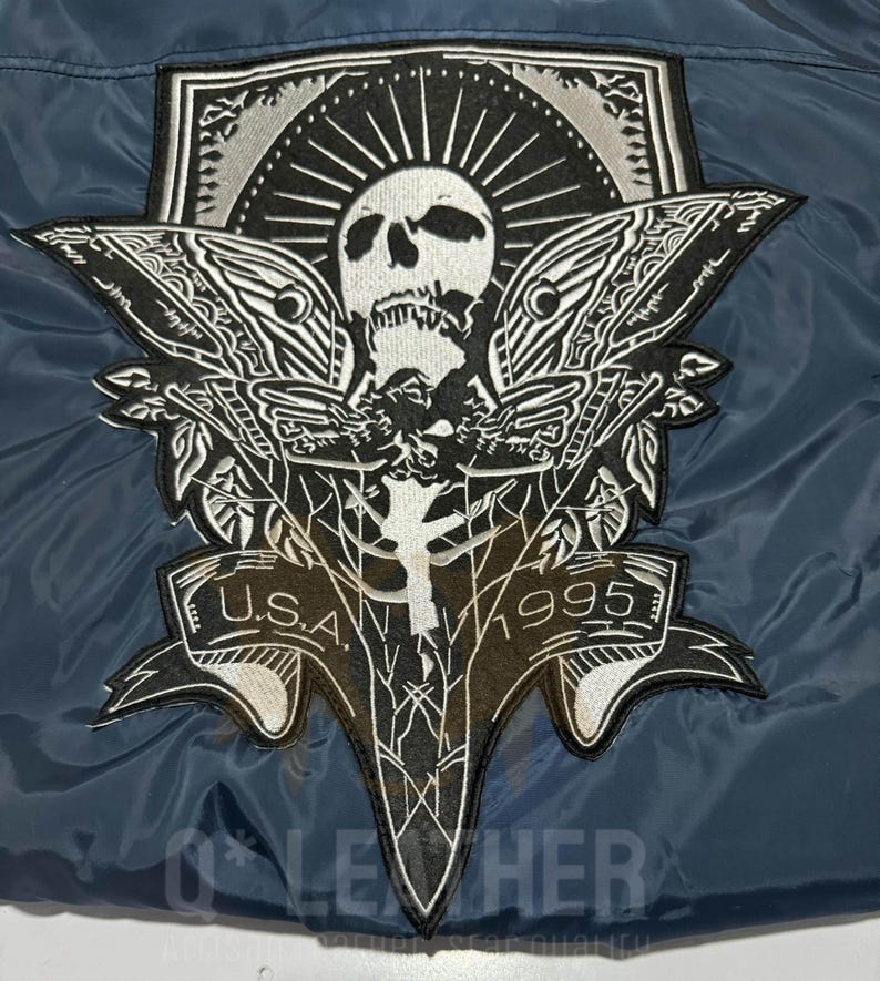 May include: A dark blue jacket featuring a large embroidered patch on the back. The patch displays a white skull with moth wings, a sunburst design, and a banner at the bottom with the text U.S.A. 1995. The embroidery uses black and white thread.