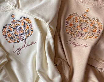 May include: Two cream and beige sweatshirts with embroidered pumpkins and names. The pumpkin is decorated with floral patterns in orange, yellow, and pink. The names are "Lydia" and "Essie".