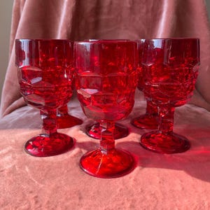 May include: A set of five ruby red wine glasses. Each glass features a faceted design on the bowl and a round base. The glasses are arranged on a pink surface, creating a visually striking display.