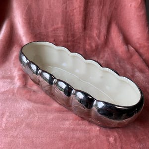 May include: A silver-colored, oblong planter with a scalloped rim and a cream-colored interior. The planter has a reflective, metallic finish and is likely made of ceramic or a similar material. The design is simple yet elegant, suitable for various plants.