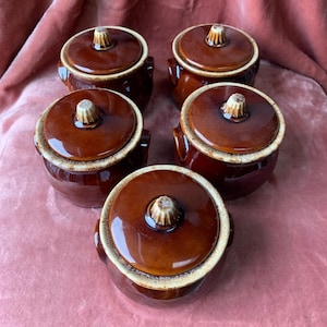 May include: Five brown ceramic lidded pots with handles. The lids have a light-colored knob on top. The pots have a glossy finish and a light-colored rim. The pots are arranged on a pink surface.