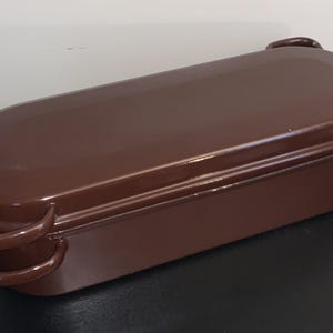May include: A brown, rectangular enamel baking dish with a lid. The dish has rounded edges and two curved handles on one side. The lid has a single handle. The dish is approximately 12 inches long.