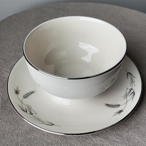 May include: A white ceramic gravy boat with a silver rim and a matching saucer. The saucer has a delicate floral design with black stems and yellow flowers.