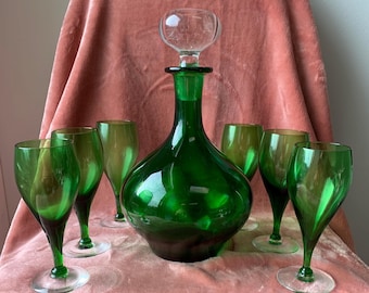 May include: A green glass decanter with a clear glass stopper, surrounded by six matching green wine glasses. The glassware has a vintage aesthetic, with a dark green hue and a rounded shape. The glasses have clear stems and bases.
