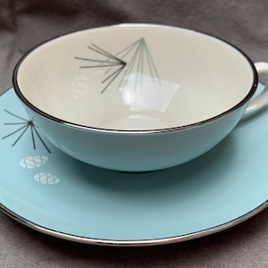 May include: A light blue teacup and saucer set with a white interior and a stylized pine cone design in black and silver. The saucer has a silver rim.
