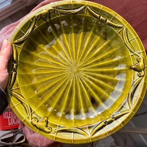 May include: A round, olive-green ceramic serving dish with a ribbed center design. The rim features decorative relief patterns and stylized embellishments. The dish is held by a hand, suggesting its size and use.