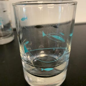 May include: Set of four clear glass drinking glasses with a turquoise fish design. The glasses are cylindrical and have a rounded bottom.