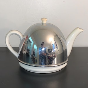 May include: A chrome-plated teapot with a white ceramic base, handle, and spout. The teapot has a rounded, dome-shaped lid with a small, cream-colored knob. The reflective surface of the teapot mirrors the surrounding environment.