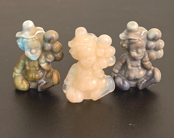 May include: Three small, seated clown figurines in varying colors. Each clown wears a hat and overalls, with a cluster of round shapes on the back. The clowns are in shades of blue, beige, and brown.