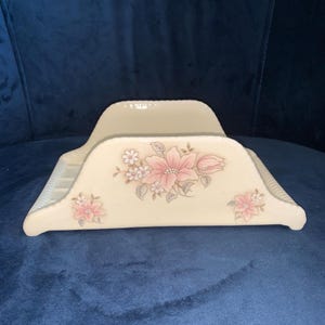 May include: Cream-colored ceramic napkin holder with a floral design. The holder features pink flowers and gray leaves. The edges have a ridged texture. The design is symmetrical, with flowers on both sides.