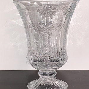 May include: A clear glass vase with a pedestal base and palm tree etching. The vase is tall and slender with a wide mouth.