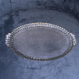 May include: An oval-shaped, clear glass tray with a textured surface and a gold-colored, ornate trim. The tray has two decorative handles on each end. The tray is designed for serving or display.