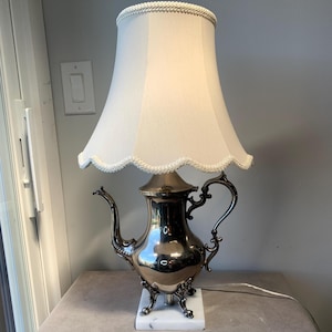 May include: A unique table lamp featuring a silver teapot base with a white lampshade. The teapot base is detailed with ornate handles and a spout, mounted on a white marble base. The lampshade is a soft white with a scalloped trim.