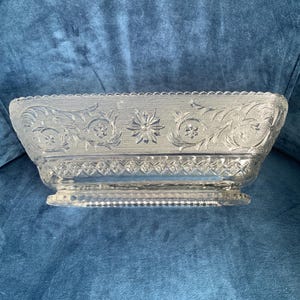 May include: A clear, rectangular glass dish with ornate floral and starburst patterns. The dish has a scalloped rim and a diamond-patterned base. The dish is raised on a small, rectangular base.