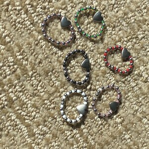 May include: Six beaded rings in various colors, including purple, green, black, red, white, and pink. Each ring features a silver heart charm. The rings are displayed on a textured, beige surface.