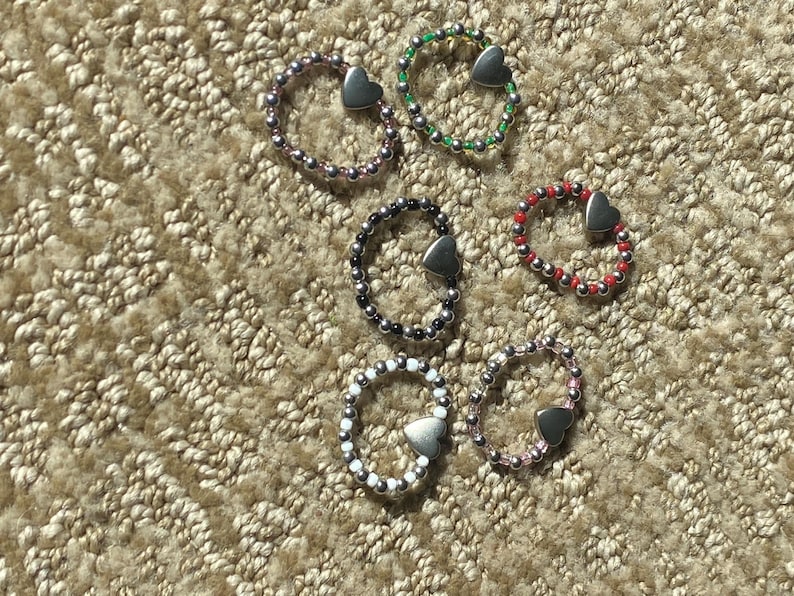 May include: Six beaded rings in various colors, including purple, green, black, red, white, and pink. Each ring features a silver heart charm. The rings are displayed on a textured, beige surface.