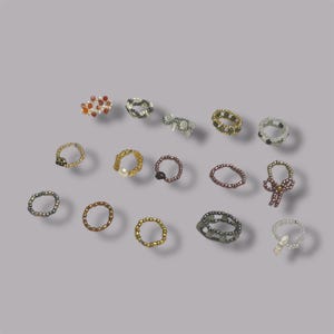 May include: A collection of beaded rings in various colors and designs. The rings feature a variety of bead sizes and colors, including gold, silver, black, white, and red. Some rings have unique embellishments, such as a pearl or a bow.