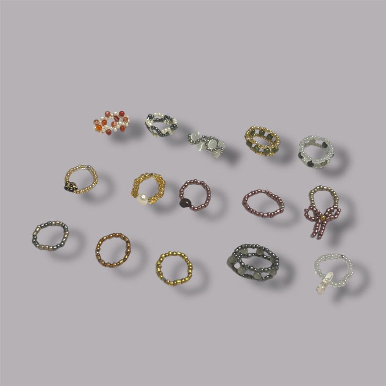May include: A collection of beaded rings in various colors and designs. The rings feature a variety of bead sizes and colors, including gold, silver, black, white, and red. Some rings have unique embellishments, such as a pearl or a bow.